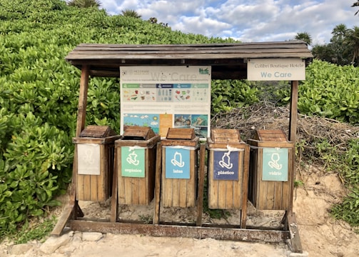 A set of five wooden recycling bins is situated outdoors on a sandy surface, surrounded by lush green foliage. Each bin is labeled for different types of waste: organic, cans, plastic, and glass. Above the bins, a wooden shelter provides protection, and there is informational signage with guidance on waste separation.